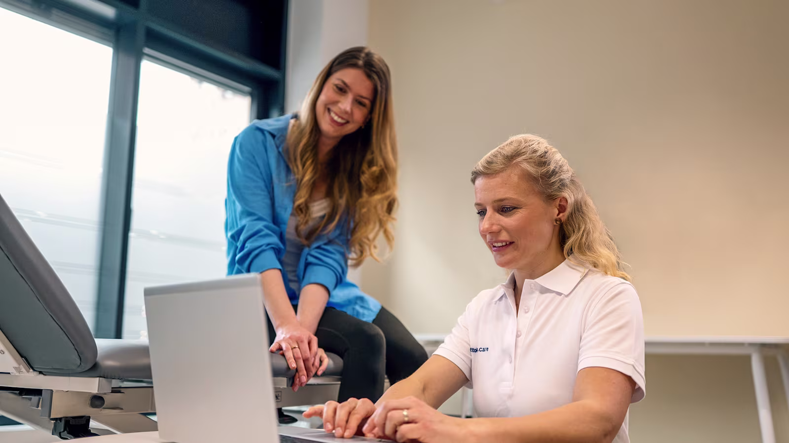 CPO tedavi odasında dizüstü bilgisayarın önünde otururken hastası gülümseyerek omzunun üzerinden bakıyor
