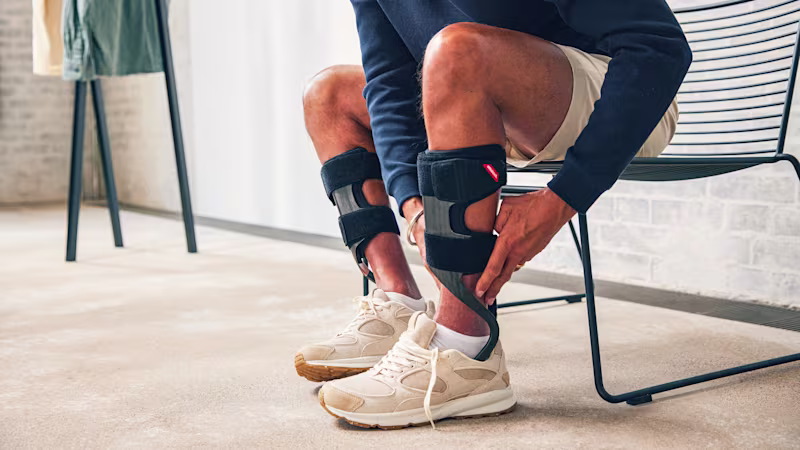 A person read as a man sits in a chair and adjusts her WalkOn Reaction lateral ankle-foot orthosis to his left leg. The image detail only shows the person's lower body.