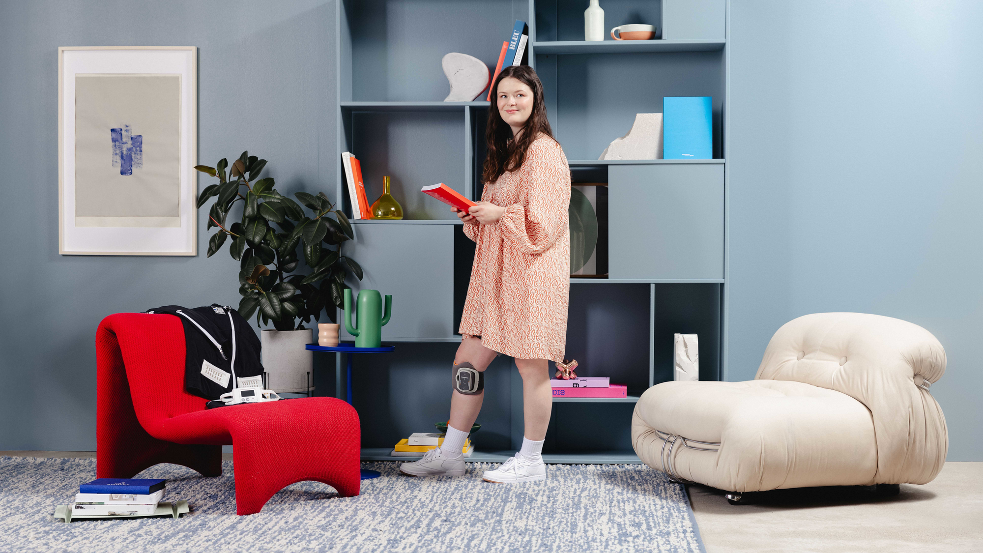 A person read as a woman is standing in a living room. She is wearing a knee-length dress and she is wearing the L300 Go orthosis below her right knee.