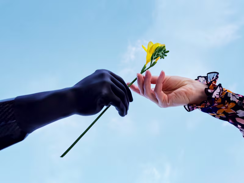 A natural hand and an Ottobock michelangelo prosthetic hand are passing a flower to each other against a sky-blue background.