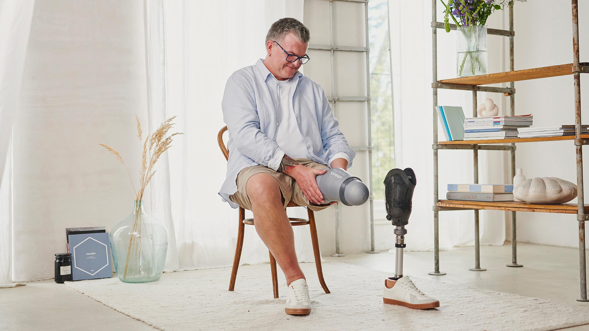 A male, transtibial user is sitting on a wodden chair, donning his liner.