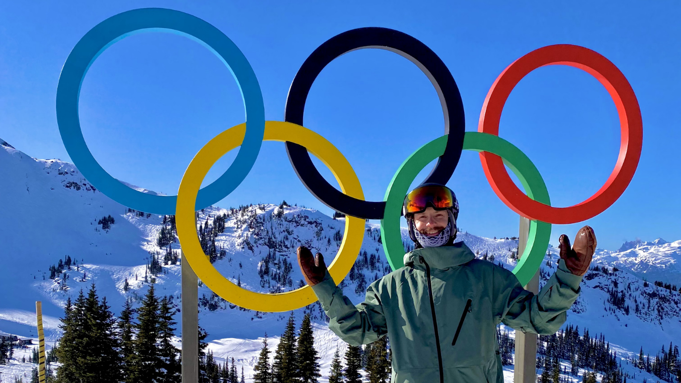 Paralympian Noah Elliot in ski gear standing happily in front of a statue of the Olympic rings with arms stretched wide 