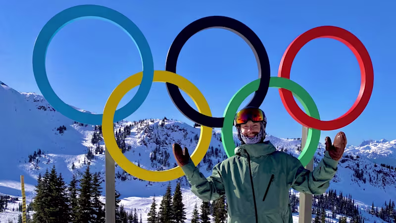 Paralympian Noah Elliot in ski gear standing happily in front of a statue of the Olympic rings with arms stretched wide