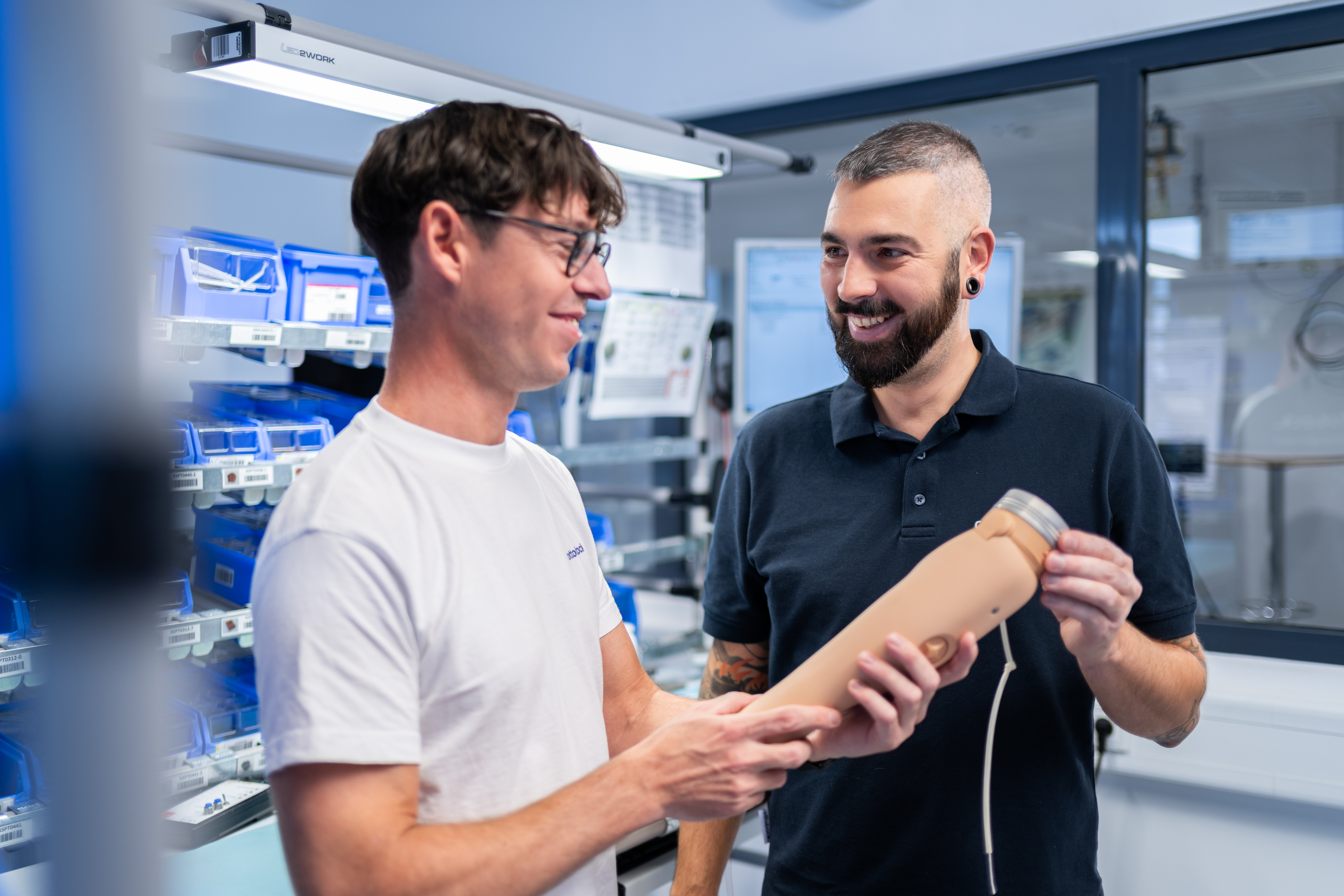 Auf dem Bild sieht man zwei Ottobock Mitarbeiter, sie lächeln beide. Zusammen halten sie eine Armprothese von Ottobock in den Händen.