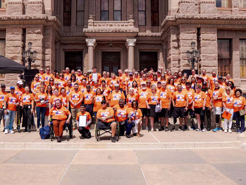 Un grupo en la coordinación de camisetas naranjas que representan el mensaje del Mes de Concientización sobre la Pérdida de Extremidades y la Diferencia de Extremidades parado frente al edificio Capitol Hill de Austin, Texas para abogar por un proyecto de ley que apoye a las personas con diferencias de extremidades