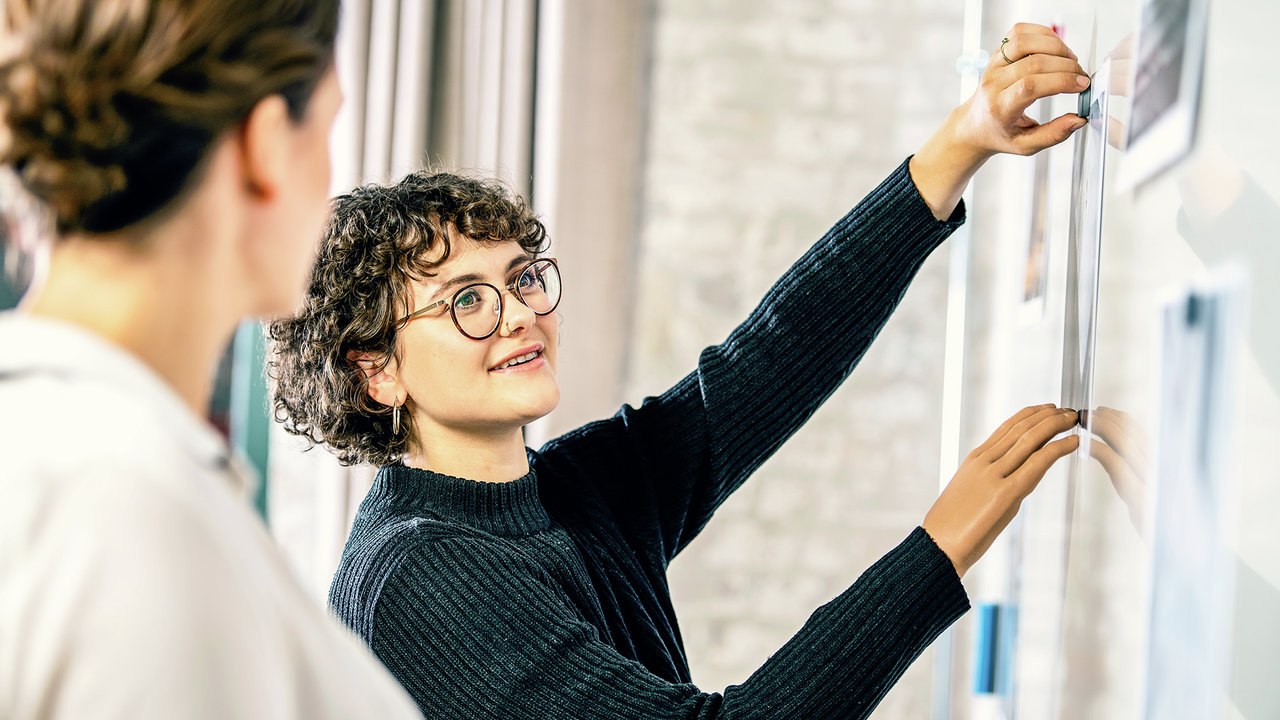 Eine Armprothesenträgerin steht vor einem Whiteboard im Büro und erklärt etwas Ihrem Kollegen. Sie trägt Myoelektrischen Armprothese mit der Myo Plus Mustererkennung von Ottobock.