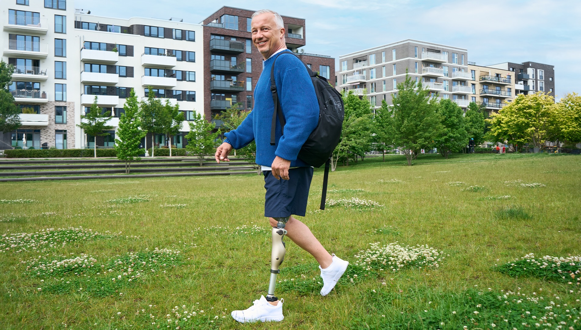 Ein amputierter Mann geht auf einer Wiese spazieren, im Hintergrund sieht man Häuser einer Wohnsiedlung. Der Mann lächelt in die Kamera und trägt das mechatronische Prothesenkniegelenk Kenevo von Ottobock.