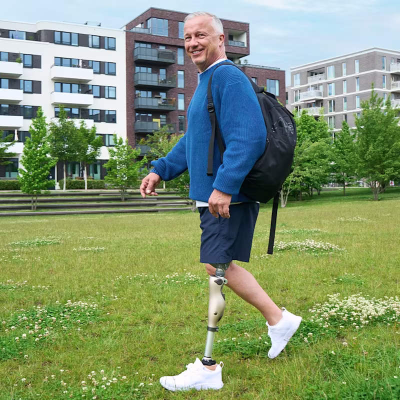 Ein amputierter Mann geht auf einer Wiese spazieren, im Hintergrund sieht man Häuser einer Wohnsiedlung. Der Mann lächelt in die Kamera und trägt das mechatronische Prothesenkniegelenk Kenevo von Ottobock.