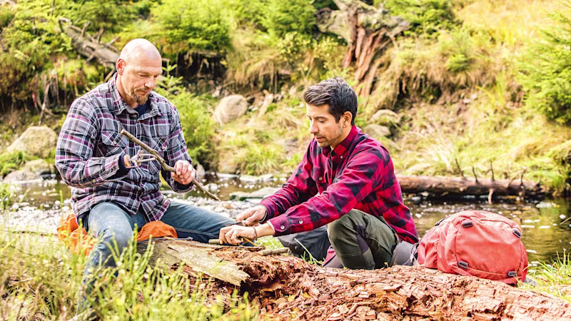 Ein Armprothesenträger sitzt im Wald auf einem Baumstumpf und schneidet mit einem anderen Mann Holz klein. Er trägt Holgers das DynamicArm-Ellbogengelenk mit der VariPlus Speed Hand von Ottobock.