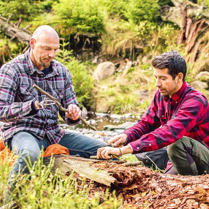 Ein Armprothesenträger sitzt im Wald auf einem Baumstumpf und schneidet mit einem anderen Mann Holz klein. Er trägt Holgers das DynamicArm-Ellbogengelenk mit der VariPlus Speed Hand von Ottobock.