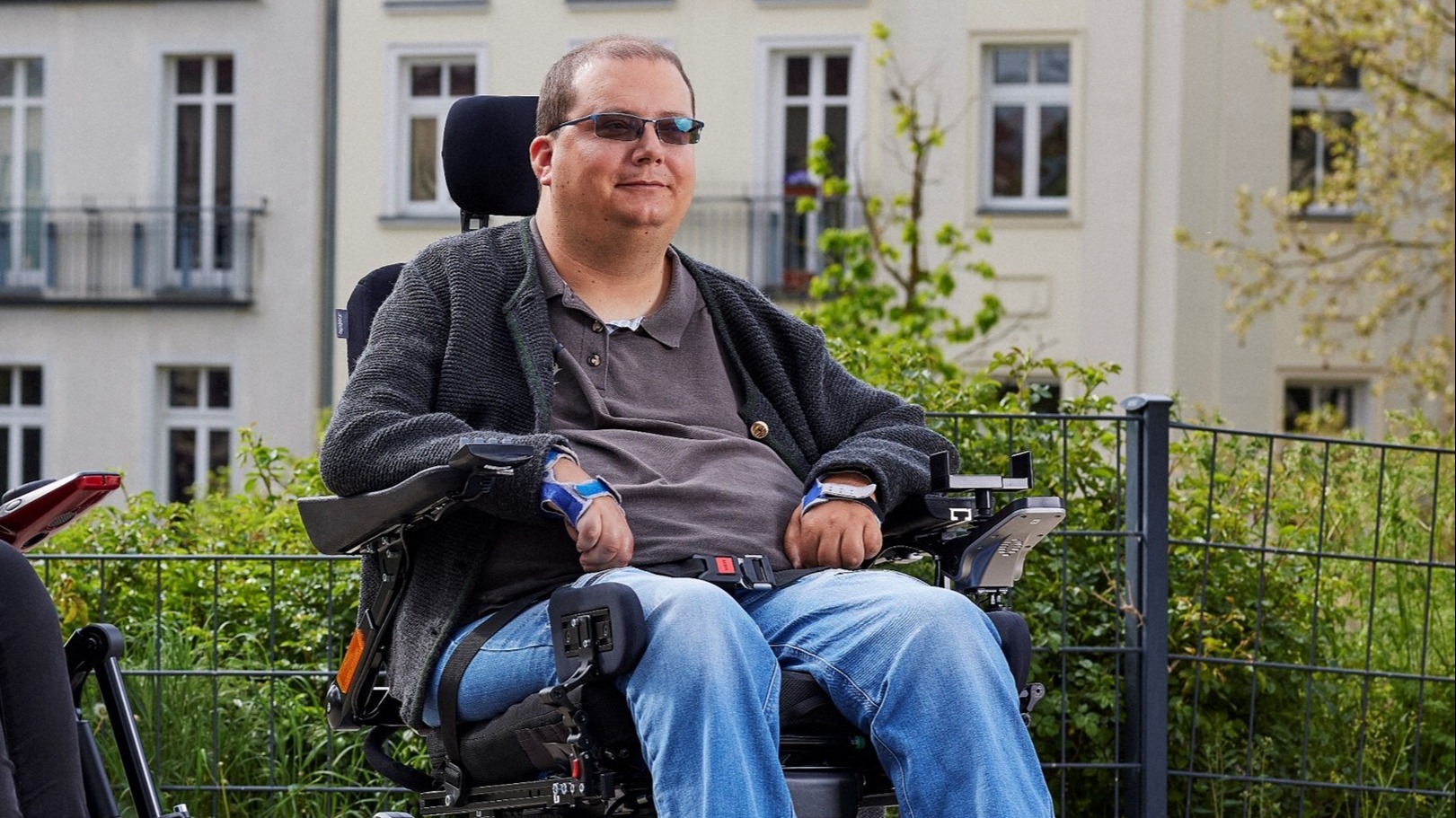 Male person with sunglasses in an Ottobock Juvo power wheelchair in front of a house facade