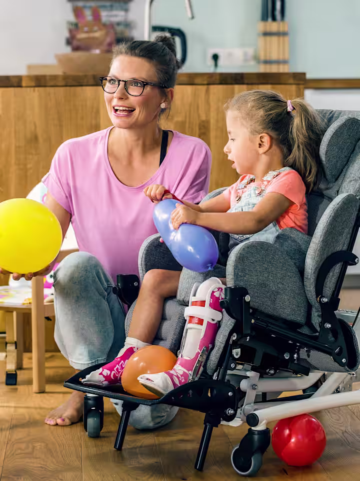 Eine Frau spielt im Wohnzimmer mit zwei Kindern. Ein Kind hat von Ottobock das Kimba Home Zimmeruntergestell.