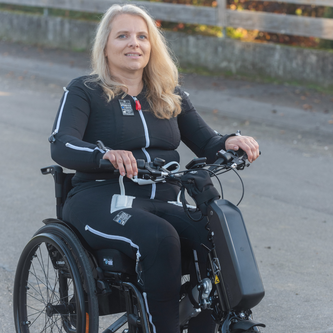 Kerstin siede su una carrozzina Avantgarde DV e utilizza un Klaxon Klick Race come dispositivo di trazione. Indossa la neurotuta Exopulse.