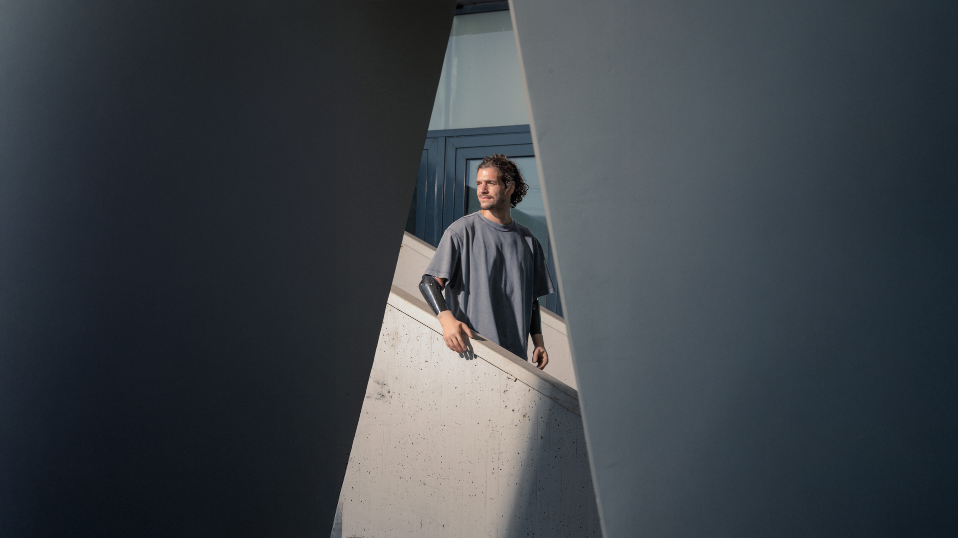 Hombre con brazos protésicos de pie entre estructuras arquitectónicas, simbolizando la resiliencia e independencia en la campaña Invisible Class de Ottobock sobre romper barreras de viaje para personas con discapacidad.