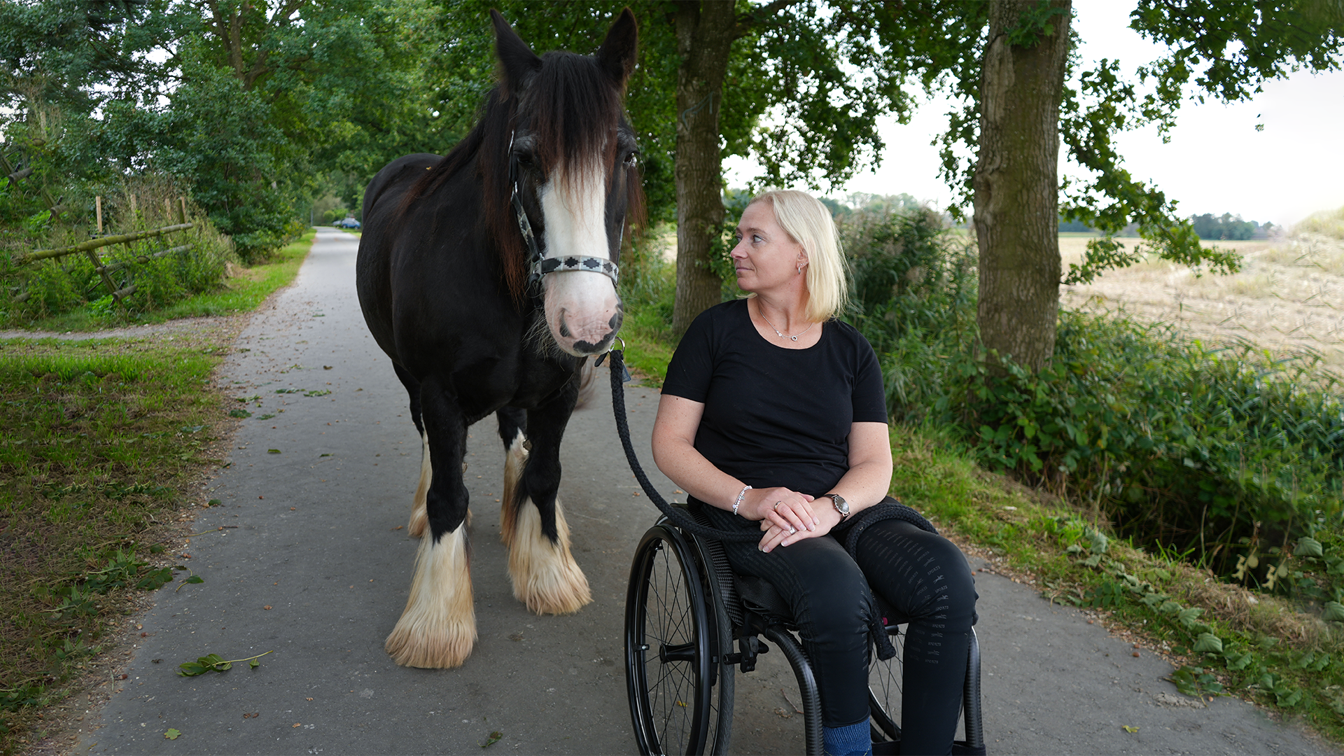 Eine weiblich gelesene Person sitzt im Zenit Rollstuhl von Ottobock. Sie steht auf einem verkehrsberuhigten Weg. Links neben der Person steht ein braunes Pferd. Die weiblich gelesene Person schaut das Pferd zuneigungsvoll an.