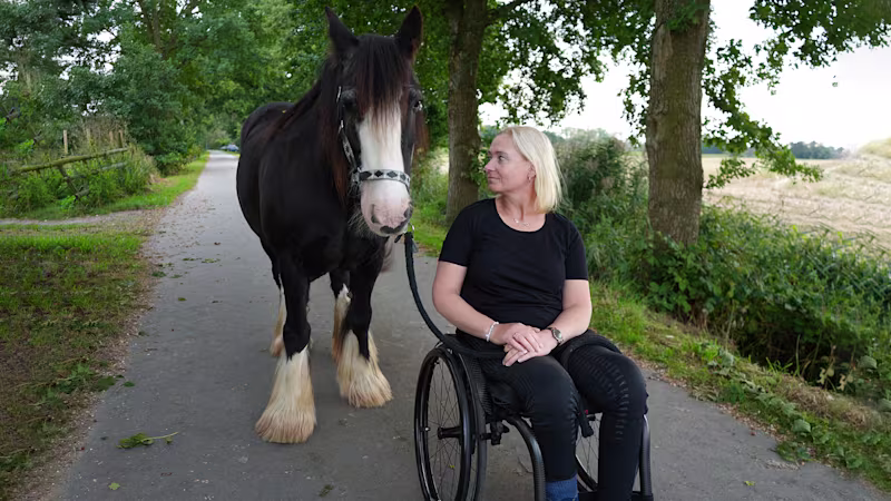 Eine weiblich gelesene Person sitzt im Zenit Rollstuhl von Ottobock. Sie steht auf einem verkehrsberuhigten Weg. Links neben der Person steht ein braunes Pferd. Die weiblich gelesene Person schaut das Pferd zuneigungsvoll an.