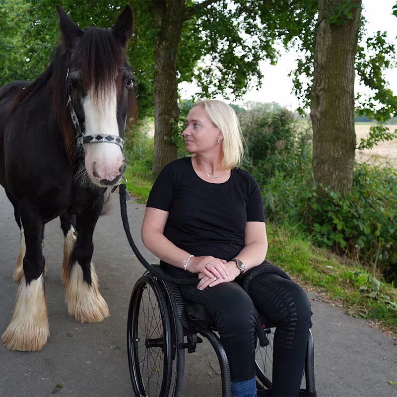 Eine weiblich gelesene Person sitzt im Zenit Rollstuhl von Ottobock. Sie steht auf einem verkehrsberuhigten Weg. Links neben der Person steht ein braunes Pferd. Die weiblich gelesene Person schaut das Pferd zuneigungsvoll an.