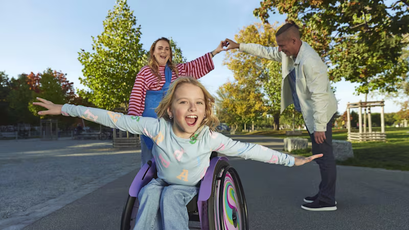 A young girl sitting in her kidevo wheelchair with her arms outstretched (as if she was flying) and her parents form a heart with their hands in the background.