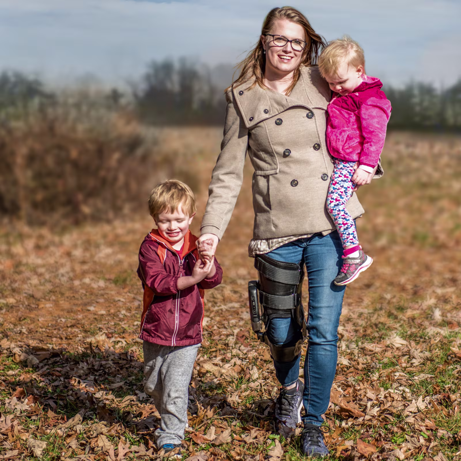 A mother holds one child while holding hands with another as they stroll outside through a field. The mother is wearing the Ottobock's C-Brace.
