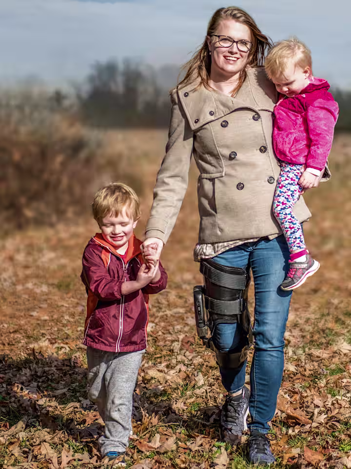 A mother holds one child while holding hands with another as they stroll outside through a field. The mother is wearing the Ottobock's C-Brace.