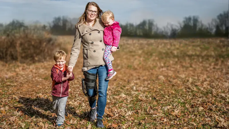 A mother holds one child while holding hands with another as they stroll outside through a field. The mother is wearing the Ottobock's C-Brace.