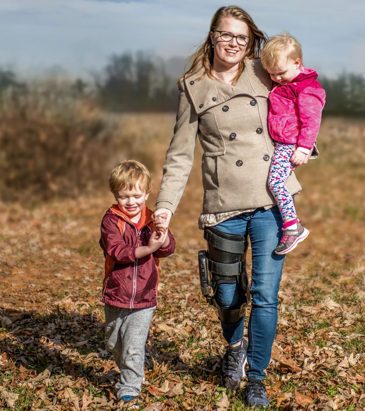 A mother holds one child while holding hands with another as they stroll outside through a field. The mother is wearing the Ottobock's C-Brace.