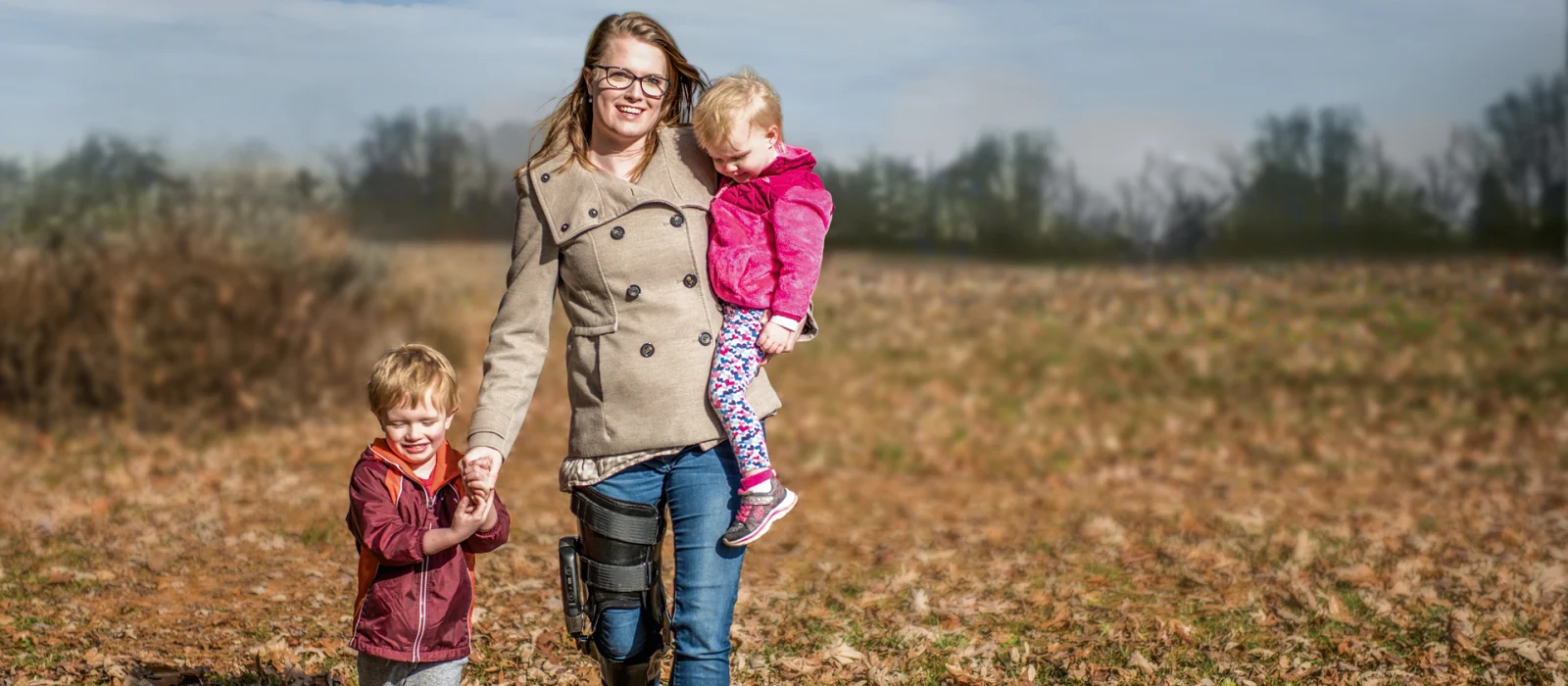 A mother holds one child while holding hands with another as they stroll outside through a field. The mother is wearing the Ottobock's C-Brace.