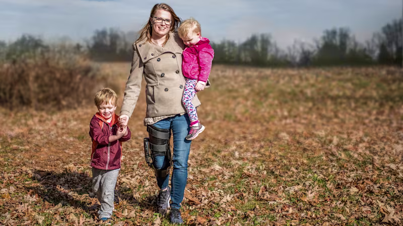 Una madre tiene in braccio un bambino mentre tiene per mano un altro mentre passeggiano fuori in un campo. La madre indossa l'ortesi C-Brace Ottobock.