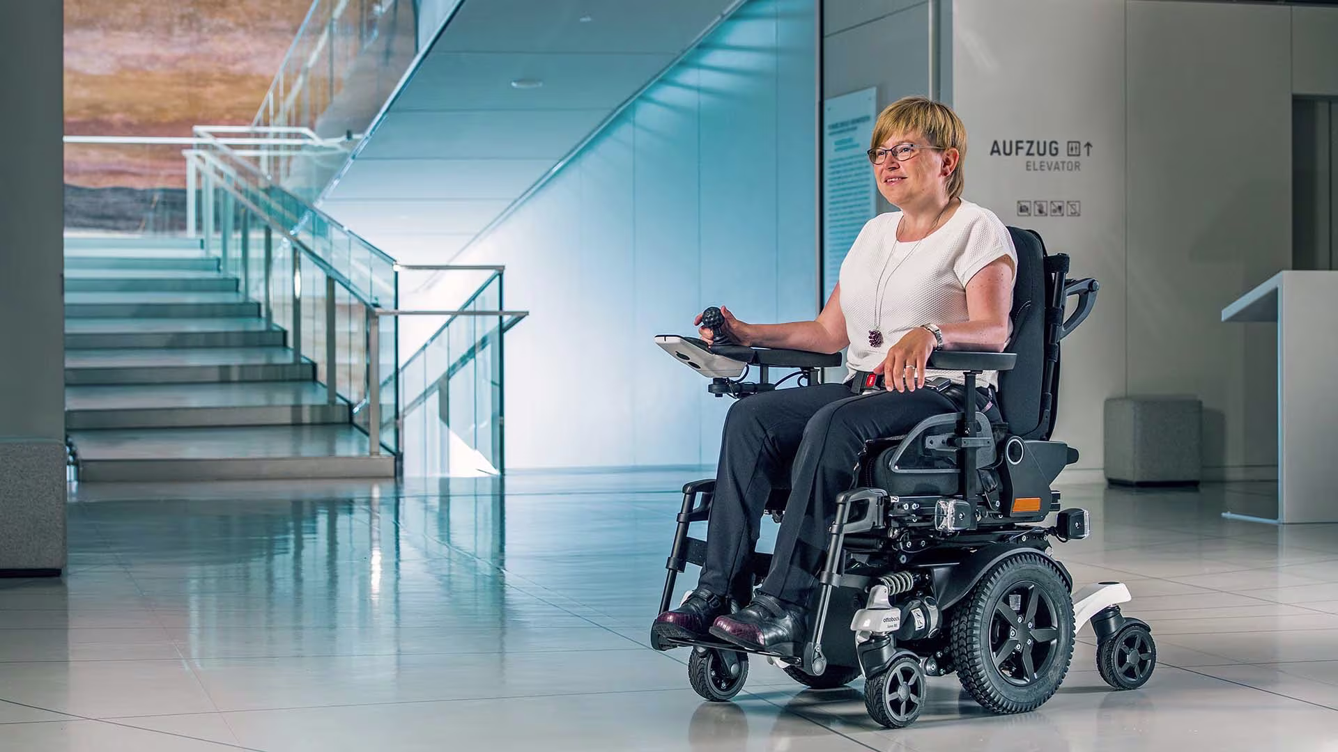 Eine Frau mit blonden kurzen Haaren sitzt im Juvo Elektrorollstuhl von Ottobock. Im Hintergrund erkennt man ein Treppenhaus eines Firmengebäudes.