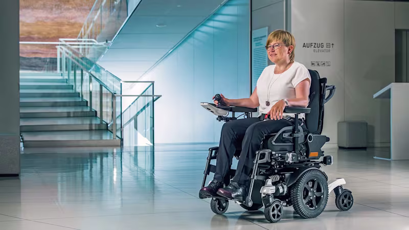 Eine Frau mit blonden kurzen Haaren sitzt im Juvo Elektrorollstuhl von Ottobock. Im Hintergrund erkennt man ein Treppenhaus eines Firmengebäudes.