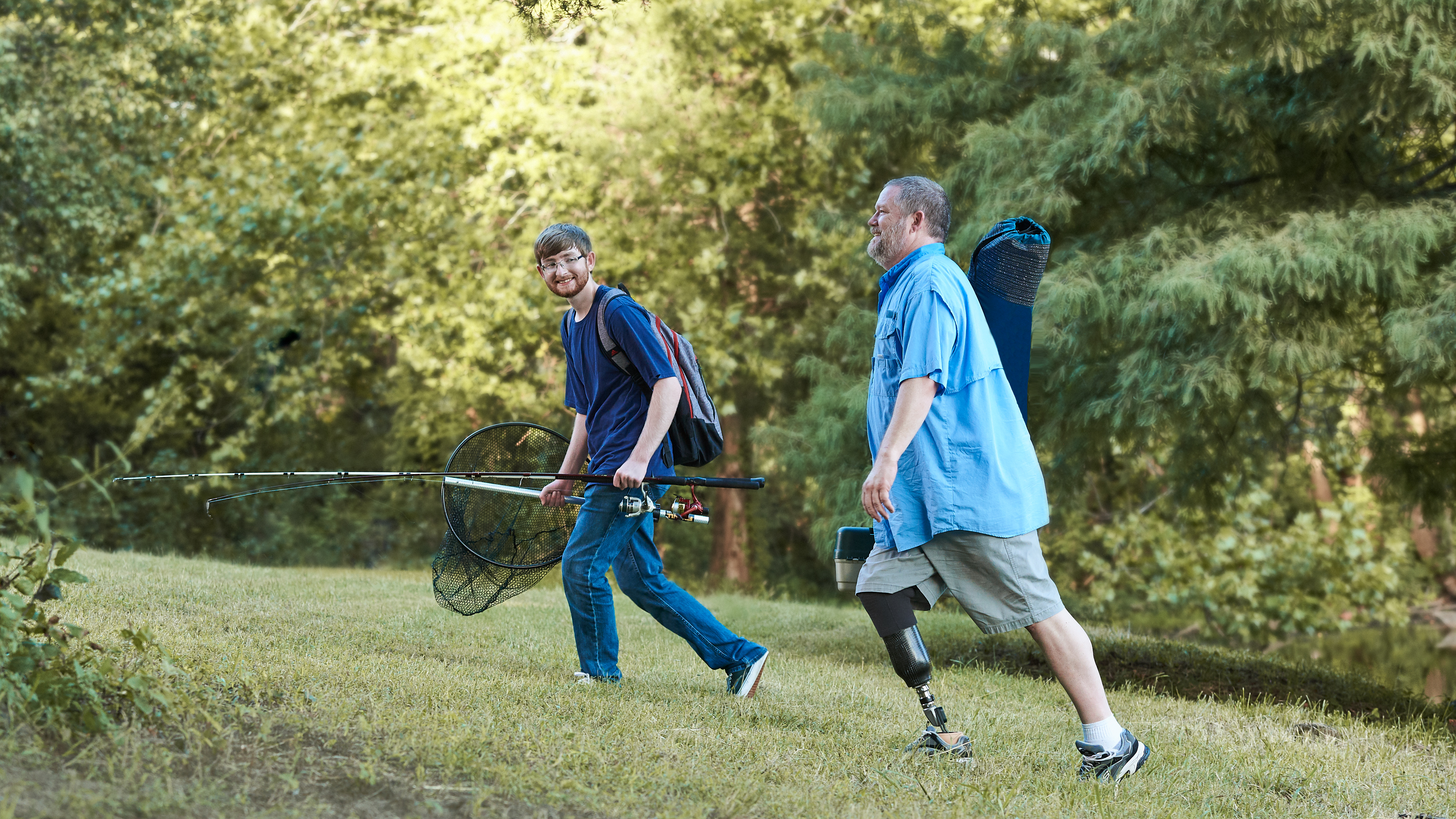 Father and son walk up a grassy hill with their fishing gear.