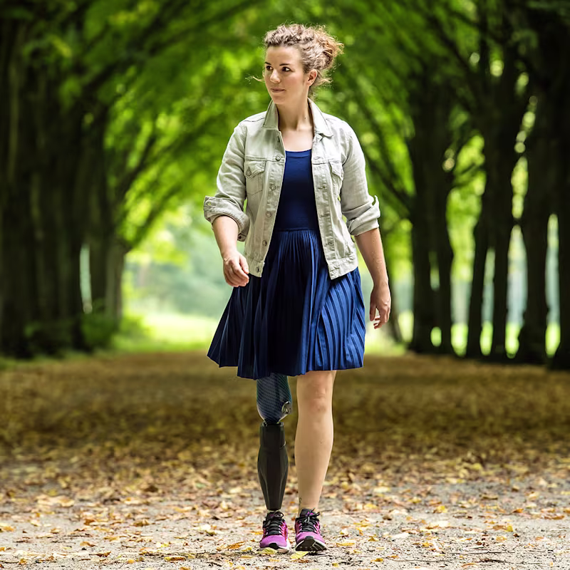 Eine weiblich gelesene Prothesenträgerin geht spazieren. Im Hintergrund ist eine Allee aus Bäumen.