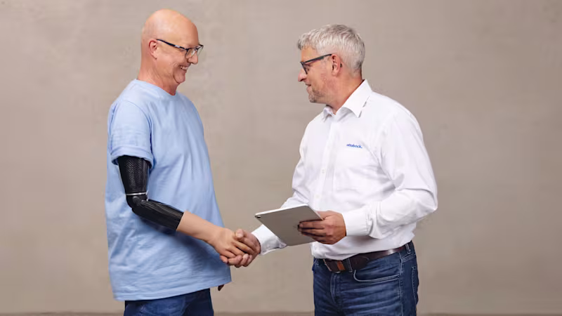 A man wearing an Ottobock speedhand upper limb prosthesis shaking hands with a certified prosthetist/orthotist in a while Ottobock shirt. The prosthetist is holding a tablet.