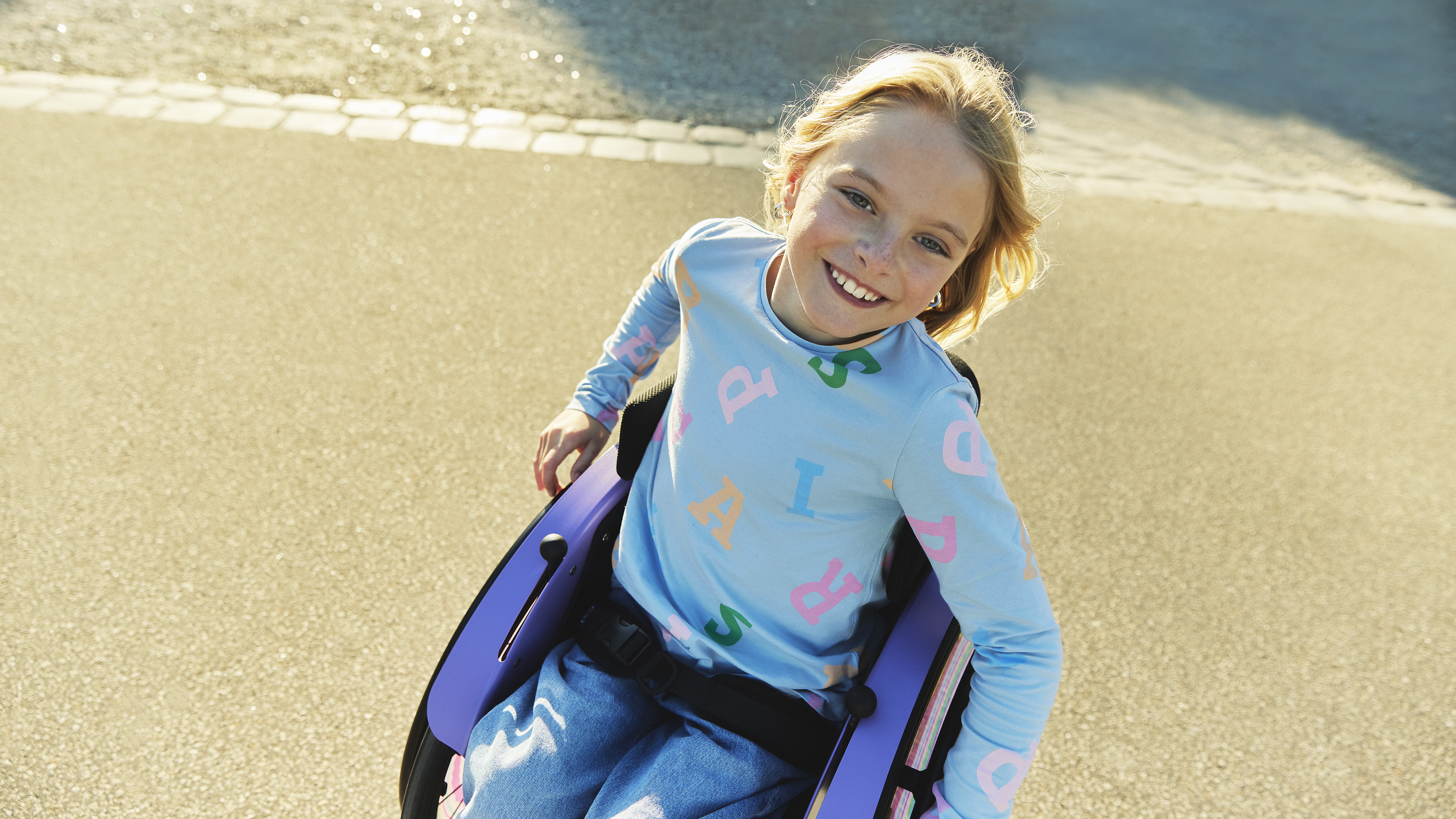 Nomine adında genç bir kız, kot pantolonu ve açık mavi gömleğiyle dışarıda kidevo tekerlekli sandalyesinde otururken genişçe gülümsüyor.