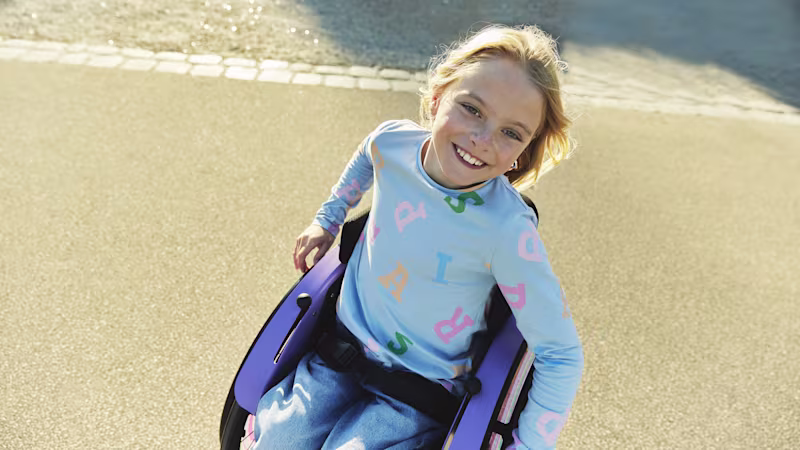 Nomine adında genç bir kız, kot pantolonu ve açık mavi gömleğiyle dışarıda kidevo tekerlekli sandalyesinde otururken genişçe gülümsüyor.