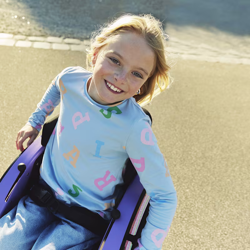 Nomine adında genç bir kız, kot pantolonu ve açık mavi gömleğiyle dışarıda kidevo tekerlekli sandalyesinde otururken genişçe gülümsüyor.