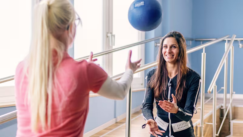 Physiotherapist and a woman in Ottobock exopulse suit at clinic, throwing a blue ball to each other.