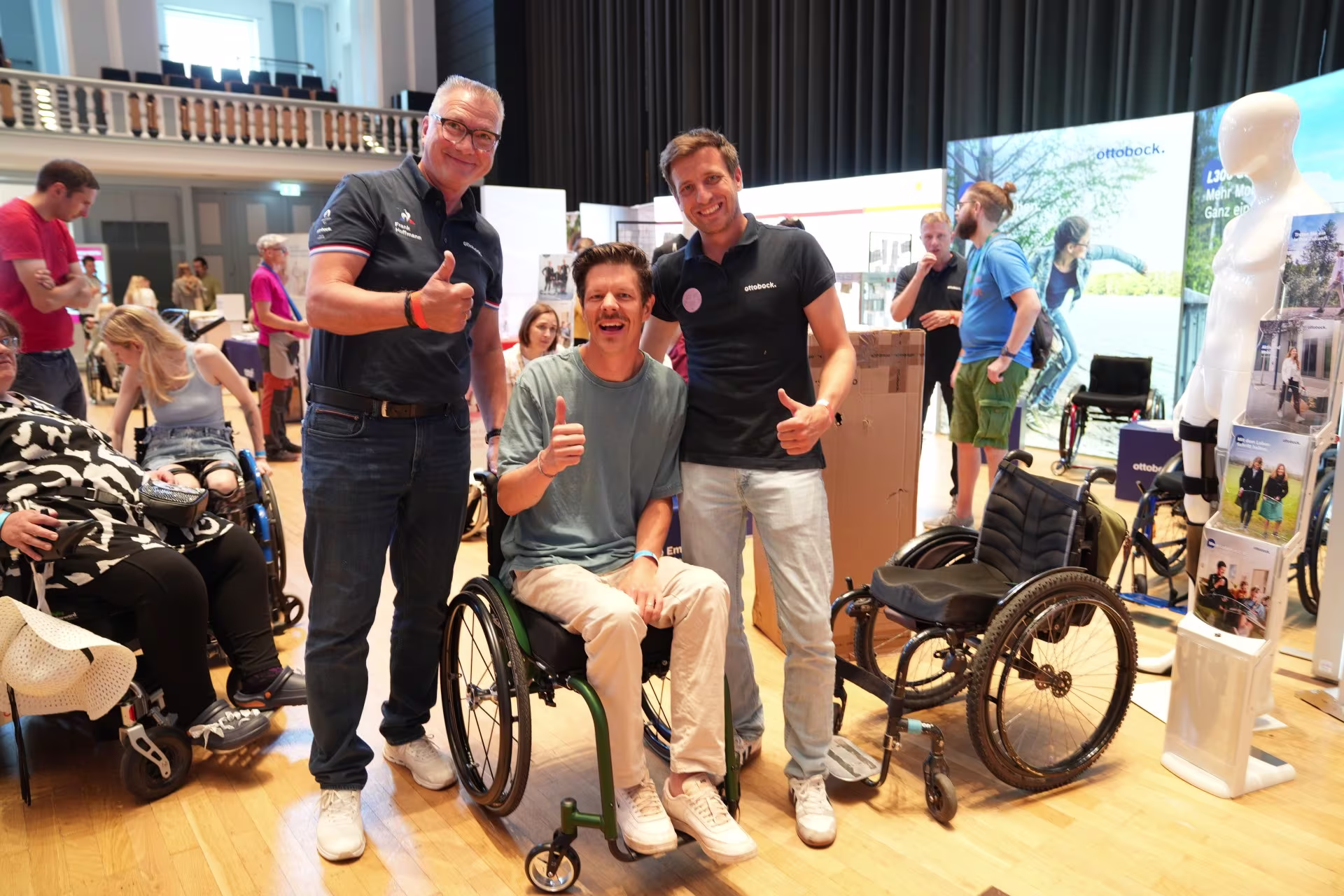 Man sieht einen jungen Mann der in einem Rollstuhl sitzt und lächelt. Rechts und links stehen Männer neben ihm, die lächelnd einen Daumen hoch zeigen.