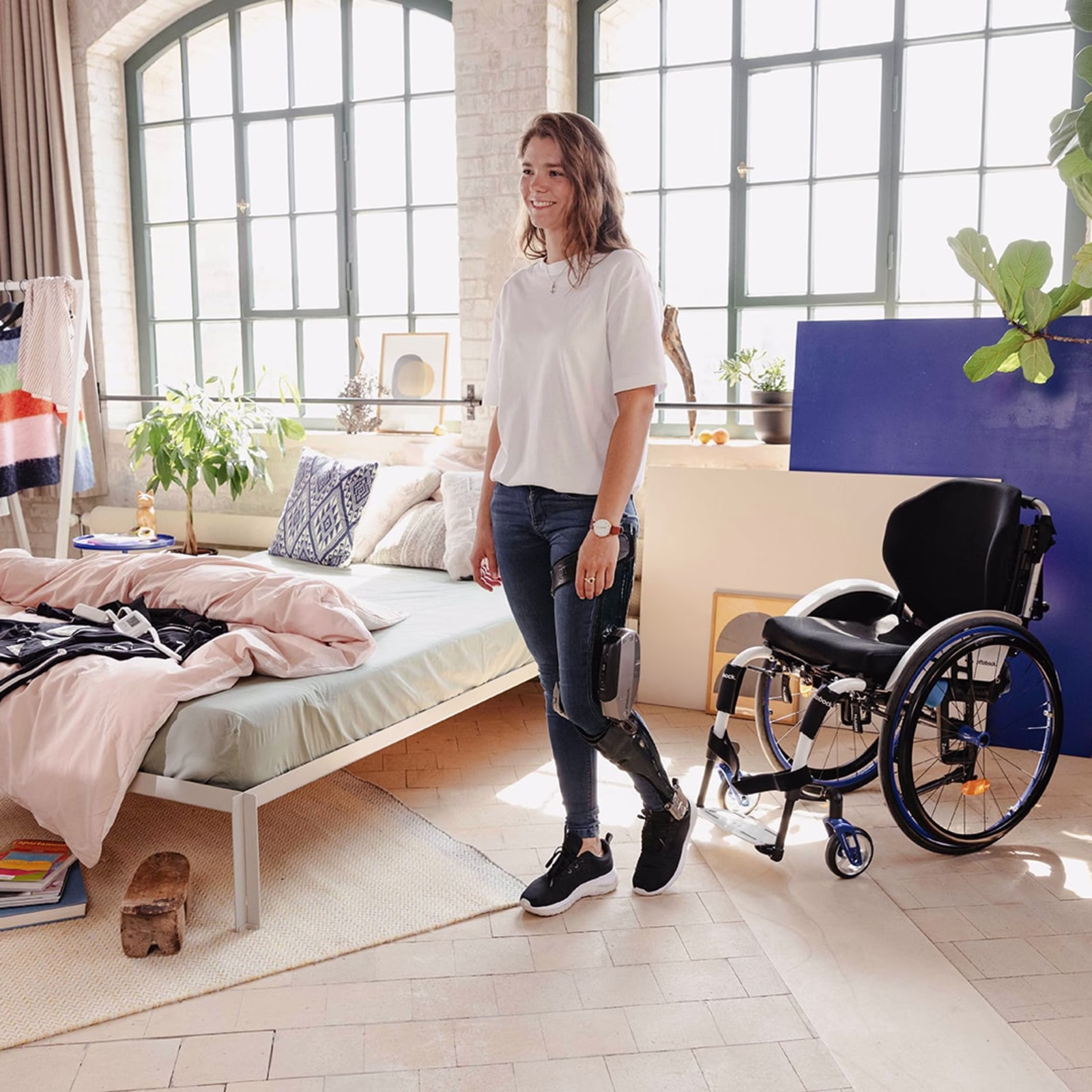 Sina, SCI patient, is wearing the C-Brace in her room. The Exopulse Mollii Suit is lying next to her as well as the wheelchair, Zenit.