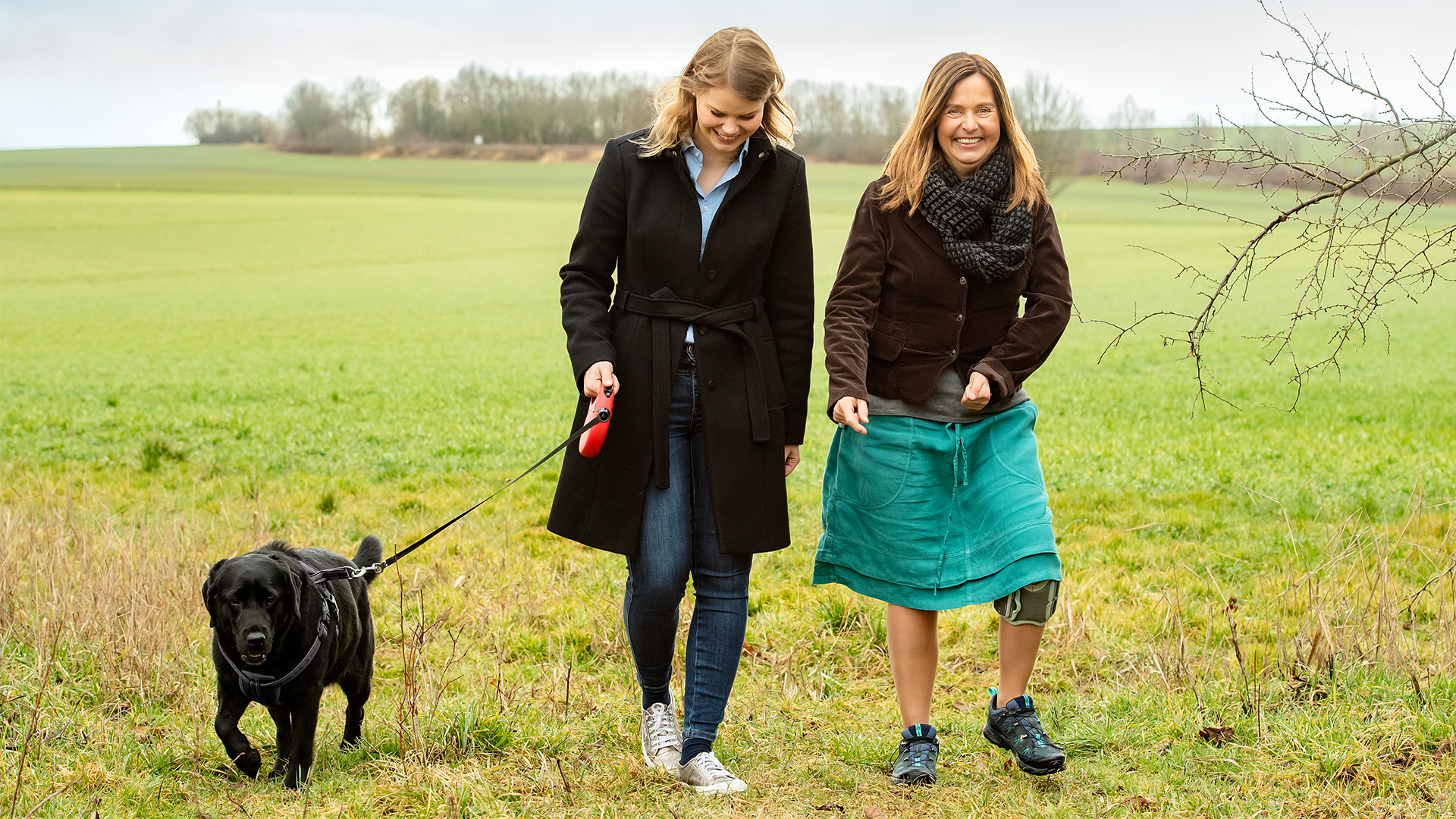 Sabine, a woman recovering from a stroke, goes for a walk in the nature accompanied by a friend with a black dog. She wears the Ottobock L300 Go.