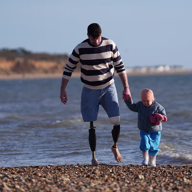 Veteran and bilateral Genium X4 user Dave Henson walks on a pebble beach holding hand with his young daughter against an ocean backdrop.