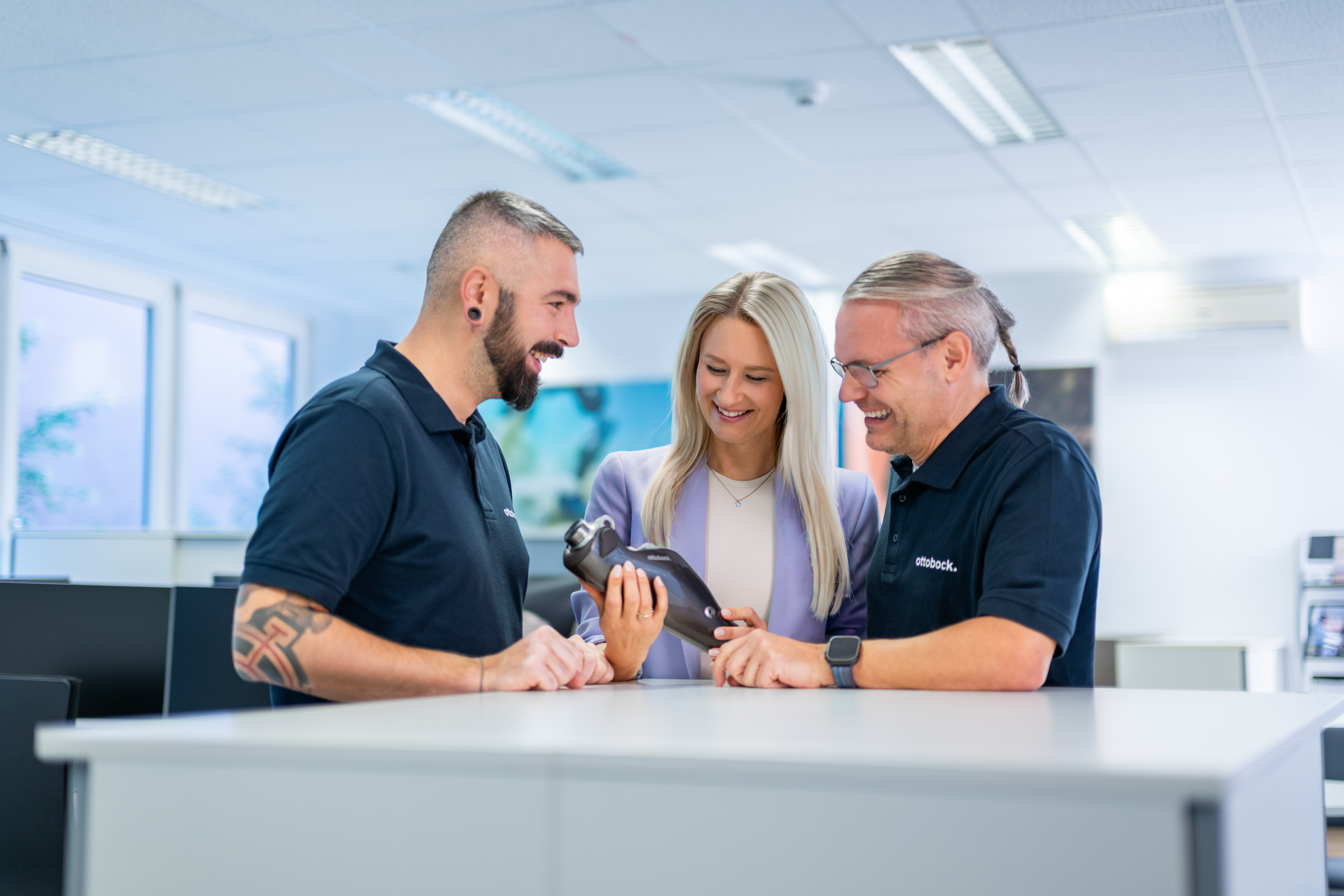 Auf dem Bild sieht man drei Ottobock Mitarbeiter. In der Mitte steht eine Frau, sie hat das C-Leg, eine Knieprothese von Ottobock in der Hand. Um sie herum stehen zwei Männer, die sich die Prothese in ihrer Hand anschauen. 