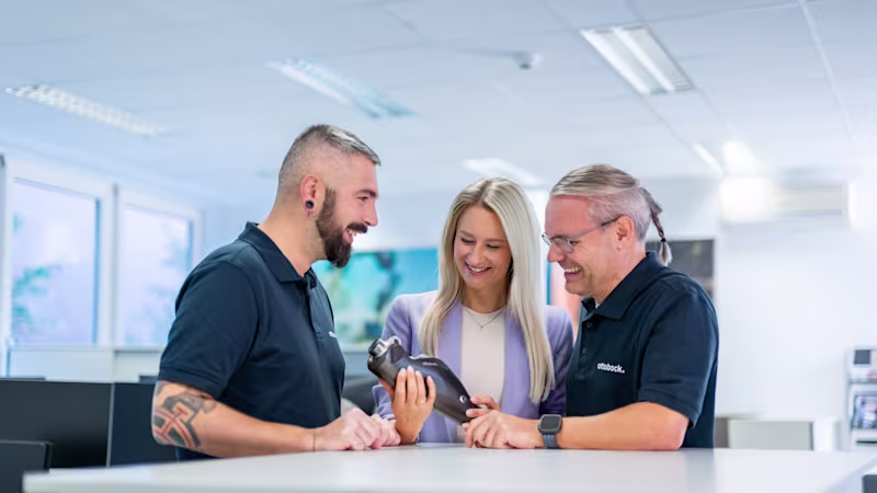 Auf dem Bild sieht man drei Ottobock Mitarbeiter. In der Mitte steht eine Frau, sie hat das C-Leg, eine Knieprothese von Ottobock in der Hand. Um sie herum stehen zwei Männer, die sich die Prothese in ihrer Hand anschauen.