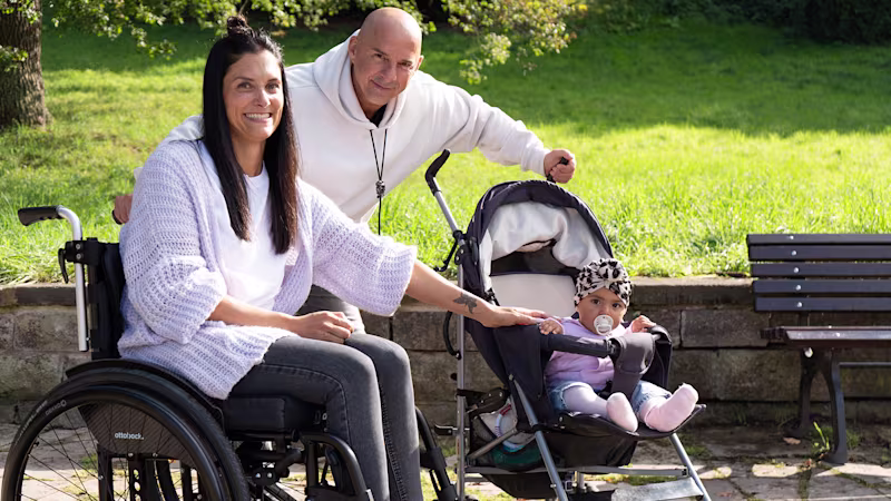 Eine Frau, ein Mann und ein Baby lächeln in die Kamera. Im Hintergrund ist ein Park zu erkennen. Die dunkelhaarige Frau sitzt im Motus Rollstuhl von Ottobock, das Baby in einem Kinder-Buggy.