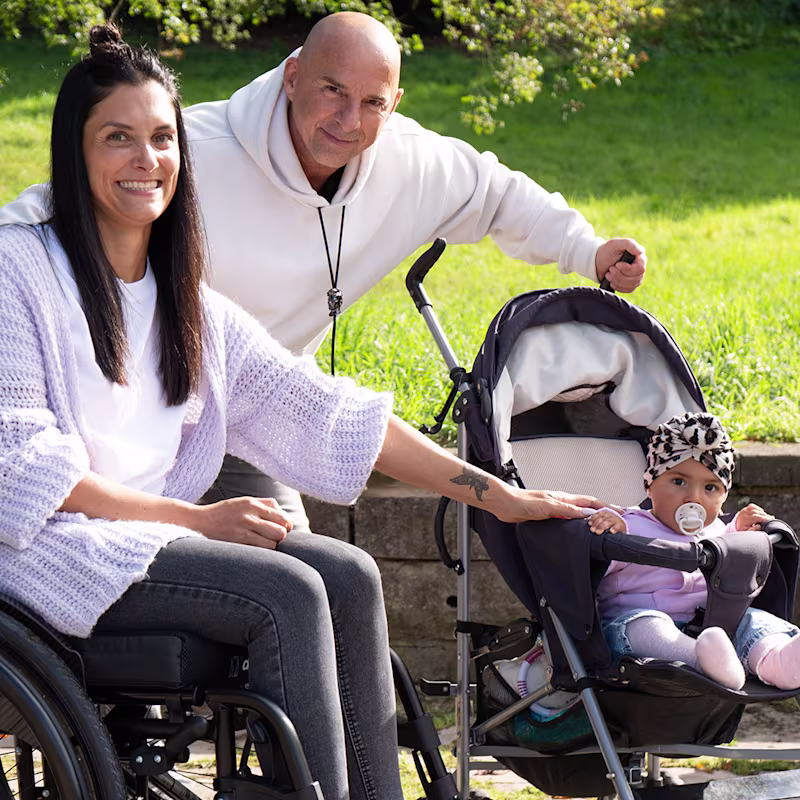 Eine Frau, ein Mann und ein Baby lächeln in die Kamera. Im Hintergrund ist ein Park zu erkennen. Die dunkelhaarige Frau sitzt im Motus Rollstuhl von Ottobock, das Baby in einem Kinder-Buggy.
