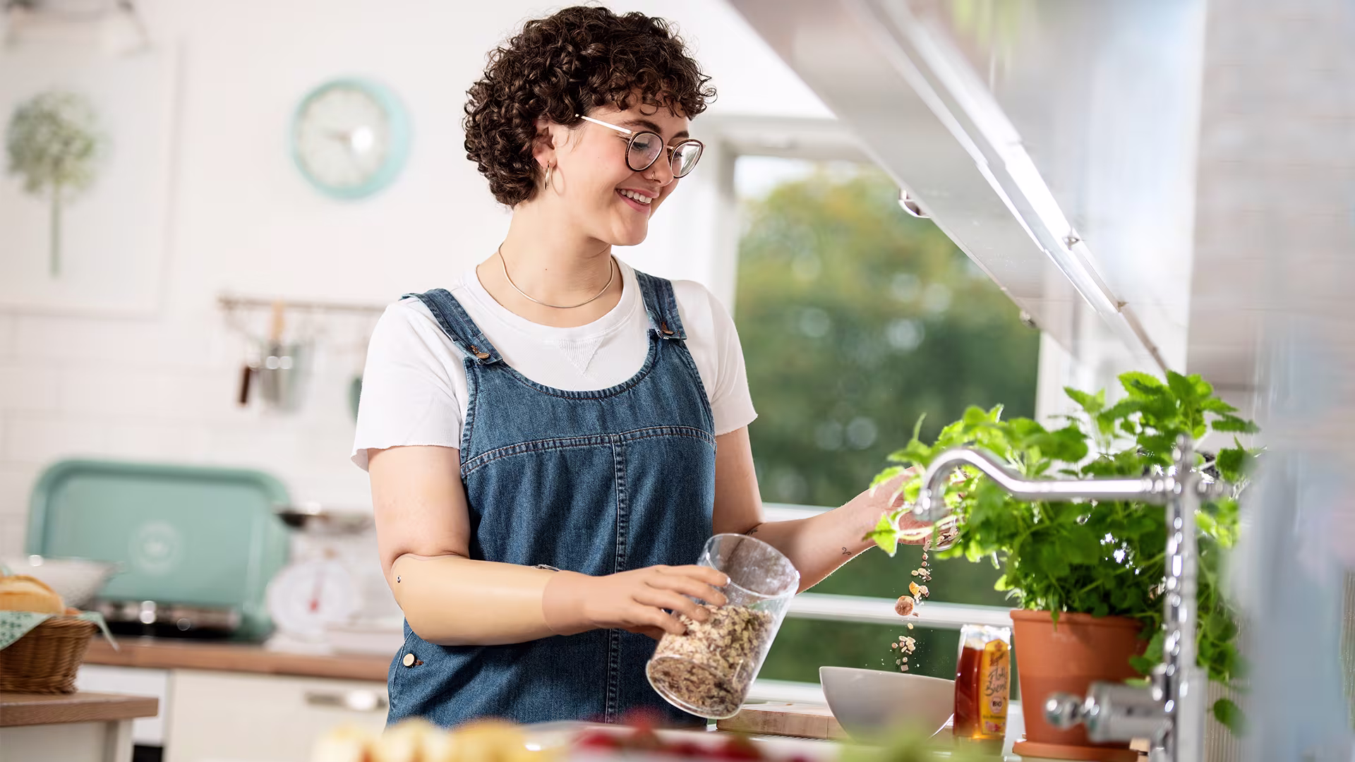Eine junge Frau steht in der Küche und bereitet Müsli zu. Dabei hält die in der linken Hand mit Hilfe ihrer bebionic Hand von Ottobock das Müsli fest.