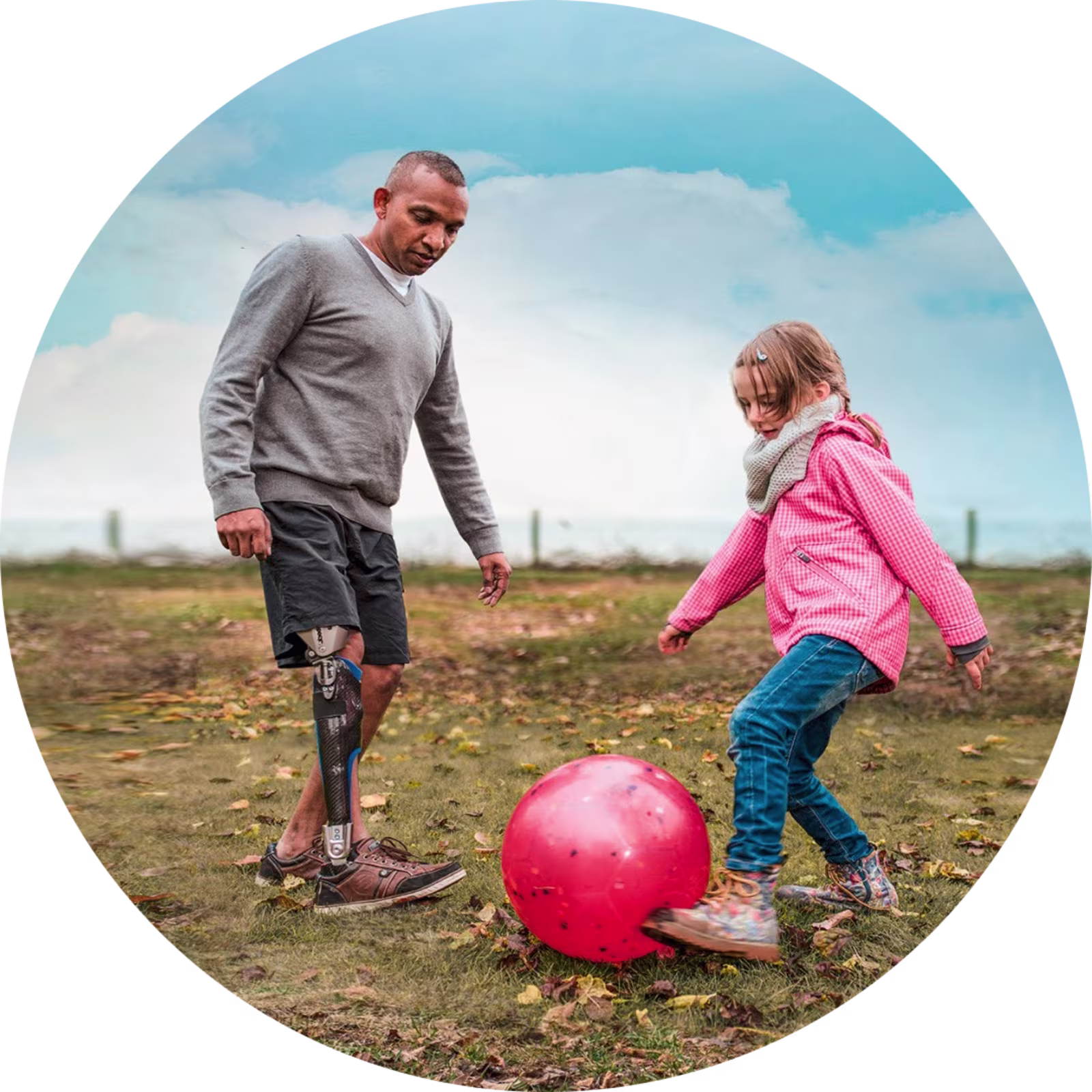 Ein männlicher Anwender spielt draußen auf dem Rasen mit seiner Tochter und einem Ball, dabei trägt er die Beinprothese C-Brace von Ottobock.