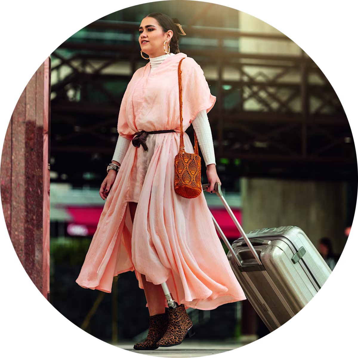 Confident woman with a prosthetic leg traveling with a suitcase, wearing a flowing pink dress and stylish accessories in an urban setting.
