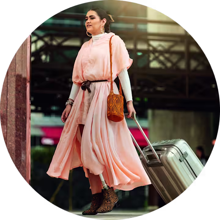 Confident woman with a prosthetic leg traveling with a suitcase, wearing a flowing pink dress and stylish accessories in an urban setting.