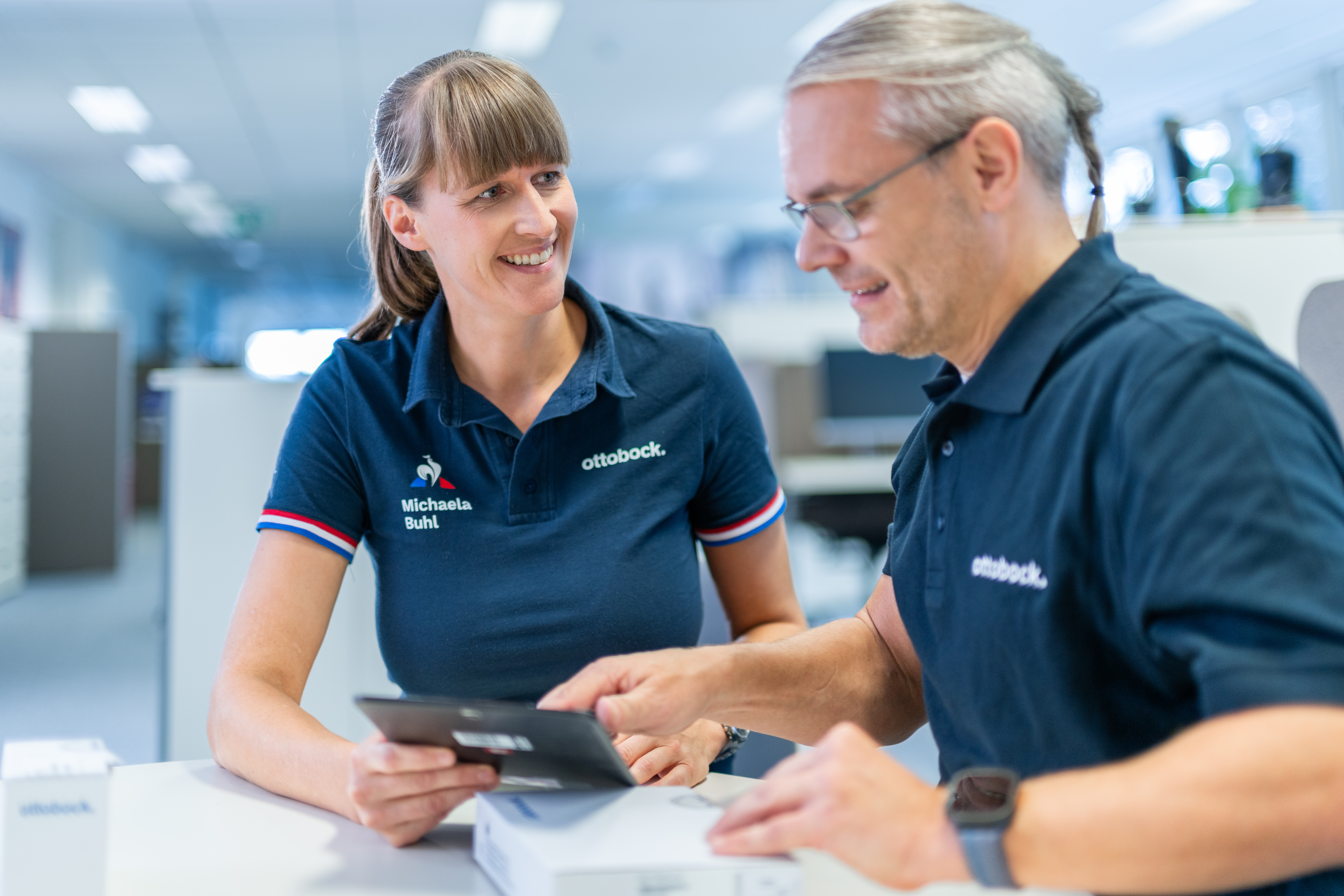 Zwei Ottobock Mitarbeiter stehen sich schräg gegenüber. Die Frau lächelt den Mann an, der Mann schaut lächelnd auf ein Tablet, welches die Frau in der Hand hält.